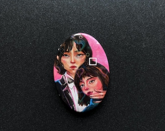 May include: Oval-shaped artwork featuring two women with dark hair and fair skin. The artwork is set against a pink background with a white square. The women are dressed in dark clothing, with one wearing a black suit jacket.
