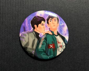 May include: Round button featuring two illustrated figures. One figure wears a green jacket with a white shirt underneath, and the other wears a gray jacket. The background is purple with white accents. The green jacket has a red X on it and the number 156.