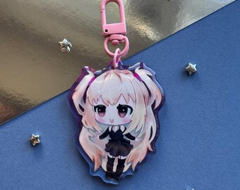 May include: A clear acrylic keychain featuring a chibi-style anime character with long blonde hair in twin tails, wearing a black dress and dark boots. The charm is attached to a light pink plastic lobster clasp and has a purple-tinted border.