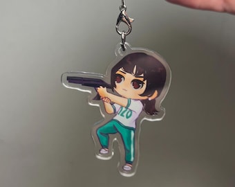 May include: Clear acrylic keychain featuring a cartoon character in a white shirt with the number "120" and teal pants, holding a black gun. The keychain has a silver clasp and is suspended against a gray background.