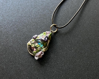 May include: A handcrafted pendant necklace featuring a teardrop-shaped pendant with a silver-tone metal frame. The pendant is adorned with colorful floral and leaf details in shades of green, purple, and pink, with iridescent accents. The necklace has a black cord.