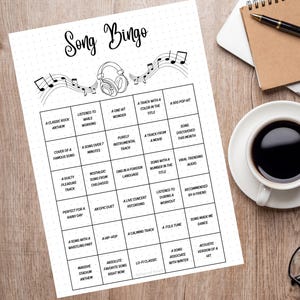 May include: A black and white "Song Bingo" game sheet with music notes and a headphone graphic. The bingo card has various music-related prompts. The sheet is on a wooden surface, next to a coffee cup, glasses, a pen, and a notebook.