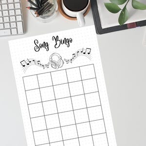 May include: A black and white Song Bingo game sheet with a grid of squares. The title "Song Bingo" is written in cursive above a musical staff with notes and headphones. The sheet is on a white surface.