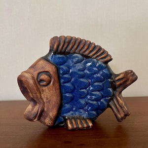 Könnte beinhalten: Eine Keramikfischfigur mit braunem Kopf, Schwanz und Flossen mit strukturierter Oberfläche. Der Körper des Fisches ist mit tiefblauen, abgerundeten Schuppen bedeckt. Der Fisch ist mit offenem Maul und geschlossenem Auge dargestellt und zeigt einen deutlichen Kontrast zwischen den braunen und blauen glasierten Bereichen des Stücks.