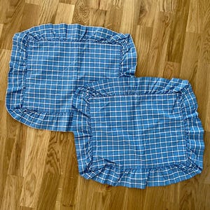May include: Two rectangular pillow shams featuring a blue and white plaid pattern. Each sham is constructed with a gathered fabric ruffle border that frames the central rectangular section, creating a consistent textile design across both pieces.