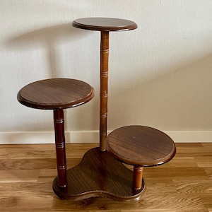 May include: A three-tiered wooden plant stand with a dark brown finish. The stand has three circular platforms of varying heights, supported by turned columns and a decorative base. The wood grain is visible.