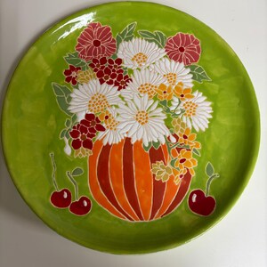 Może przedstawiać: Okrągły talerz ceramiczny z jasnym, limonkowym tłem. W centrum znajduje się wypukły, malowany wzór przedstawiający pasiastą pomarańczową dynię, która służy jako wazon na bukiet białych stokrotek, czerwonych kwiatów i żółtych pąków, z czerwonymi wiśniami.