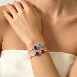 Puede incluir: Cuatro pulseras delicadas con piedras preciosas ovaladas en tonos azules, rojos y celeste. Las pulseras son de color oro y plata. Las piedras preciosas están engastadas en un diseño minimalista, mostrando los colores vibrantes de las piedras.