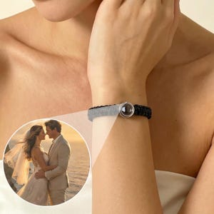 Puede incluir: Una pulsera trenzada negra y gris con un dije circular plateado. La pulsera se lleva en la muñeca. Una pequeña imagen circular de una pareja es visible en el fondo.