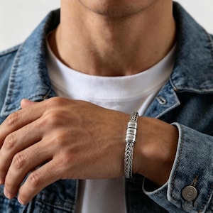 Puede incluir: Una pulsera plateada trenzada con tres pequeños dijes rectangulares grabados con los nombres "DAVID", "EMILY" y "CHLOE". La pulsera se lleva en una muñeca, sobre un fondo de chaqueta vaquera y camiseta blanca.