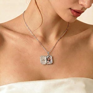 Puede incluir: Un collar con un colgante de medallón plateado rectangular. El colgante se abre para revelar una pequeña fotografía y el texto manuscrito "I love you more". El collar se lleva alrededor del cuello.