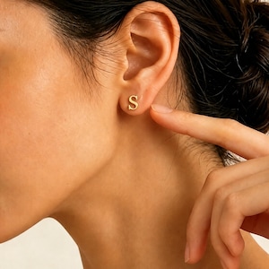 May include: A small gold stud earring shaped like the capital letter S. The earring is positioned in the earlobe, featuring a smooth, polished metallic finish and a classic serif font style that sits flush against the skin of the ear.