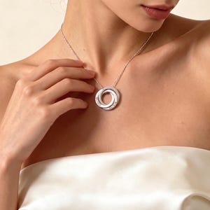 Puede incluir: Un collar plateado con tres anillos entrelazados. Cada anillo está grabado con el nombre "OLIVIA". El collar cuelga de una cadena fina. La persona lleva un top blanco sin tirantes.