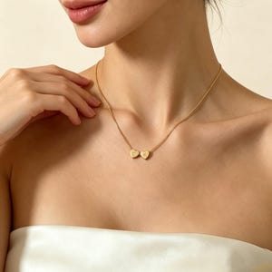 Puede incluir: Un collar dorado con dos colgantes en forma de corazón, cada uno con una inicial grabada. El collar se lleva alrededor del cuello. Los colgantes son dorados y la cadena es delicada.