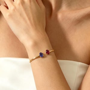 Puede incluir: Un brazalete dorado con una piedra preciosa azul y otra roja. El brazalete abierto presenta una piedra azul ovalada y una roja en cada extremo. Las piedras preciosas están pulidas.