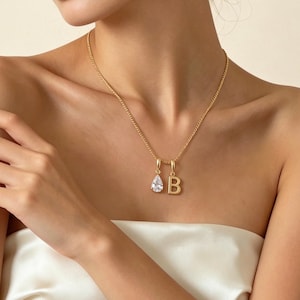 Puede incluir: Un collar dorado con una cadena y dos colgantes. Un colgante es una piedra preciosa transparente en forma de lágrima, y el otro es una letra "B" dorada. El collar se lleva alrededor del cuello.