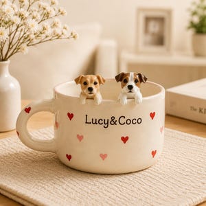 Puede incluir: Una taza de cerámica color crema con pequeños corazones rojos y rosas, el texto "Lucy&Coco" en negro y dos figuras de perros esculpidas asomándose por el borde. Un perro es beige y blanco, el otro es marrón, blanco y negro.