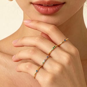 Puede incluir: Cuatro anillos de oro con gemas coloridas se muestran en una mano. Los anillos presentan pequeñas piedras vibrantes en azul, verde y una gama de colores. Los anillos son delicados y elegantes, adecuados para el uso diario.