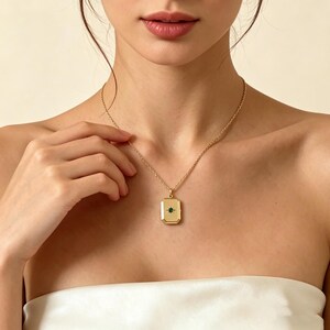 Puede incluir: Un collar dorado con un colgante rectangular. El colgante presenta una pequeña piedra preciosa en forma de estrella verde oscuro en el centro. El collar se lleva alrededor del cuello.