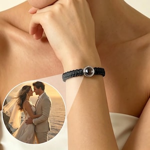 Puede incluir: Pulsera trenzada negra con un cierre plateado y una pieza central oscura y redonda. La pulsera se lleva en la muñeca. Una imagen circular de una pareja con ropa de boda está en el fondo.