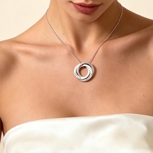 Puede incluir: Un collar plateado con tres anillos entrelazados, cada uno grabado con un nombre. El colgante cuelga de una fina cadena. La joya se muestra sobre un fondo neutro.