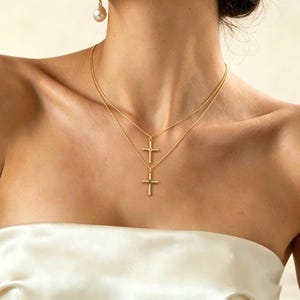 Puede incluir: Dos collares de cruz dorados superpuestos en el cuello de una persona. Los collares tienen cadenas delicadas y pequeños colgantes de cruz. Se ve un solo pendiente de perla. La persona lleva un top blanco sin tirantes.