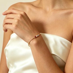Puede incluir: Una pulsera dorada con una pieza central rectangular con detalles en rojo, blanco y azul. La pulsera se lleva en la muñeca y el fondo es una tela de satén blanca.