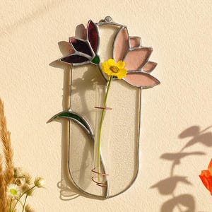 May include: A wall-mounted stained glass flower vase featuring a vertical oval frame with a clear glass test tube holder. The design includes decorative glass petals in shades of dark purple and light pink, with a single yellow flower.
