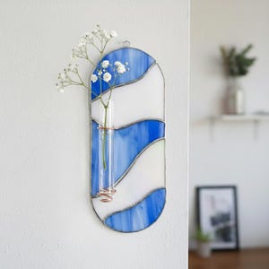May include: A stained glass wall vase with a blue and white wave design. A small glass tube holds white flowers, secured by copper wire. The vase is mounted on a white wall.