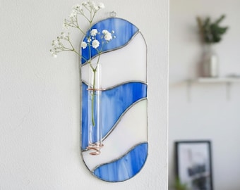 May include: A stained glass wall vase with a blue and white wave design. A small glass tube holds white flowers, secured by copper wire. The vase is mounted on a white wall.