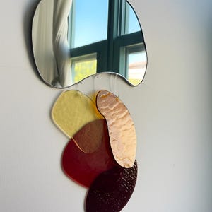 May include: A contemporary wall art piece. A wavy-edged mirror is positioned above a cluster of colourful, irregularly shaped acrylic pieces. The acrylic elements are in shades of yellow, red, and brown, creating a layered, abstract design. The mirror reflects a window.