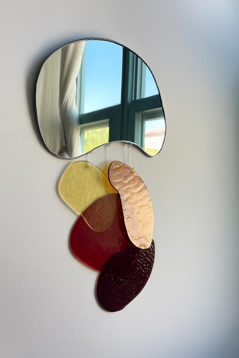 May include: A contemporary wall art piece. A wavy-edged mirror is positioned above a cluster of colourful, irregularly shaped acrylic pieces. The acrylic elements are in shades of yellow, red, and brown, creating a layered, abstract design. The mirror reflects a window.