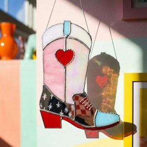 May include: A colourful stained glass cowboy boot decoration. The boot features a red heart, stars, and a checkered pattern. It is made of pink, white, red, black, and blue glass, and is suspended by a silver chain. A decorative piece.