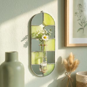 May include: A stained glass wall vase with a clear glass tube for flowers. The vase has a green and mirrored glass design, with a metal frame. A daisy and other small white flowers are in the vase. The vase is hanging on a light green wall.