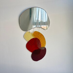 May include: A wall-mounted mirror with an unusual, freeform design. The silver-toned mirror hangs above a collection of overlapping, colourful, abstract shapes in yellow, orange, red, and burgundy. The shapes appear to be made of translucent glass.
