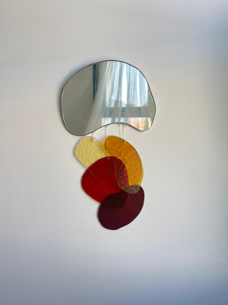 May include: A wall-mounted mirror with an unusual, freeform design. The silver-toned mirror hangs above a collection of overlapping, colourful, abstract shapes in yellow, orange, red, and burgundy. The shapes appear to be made of translucent glass.