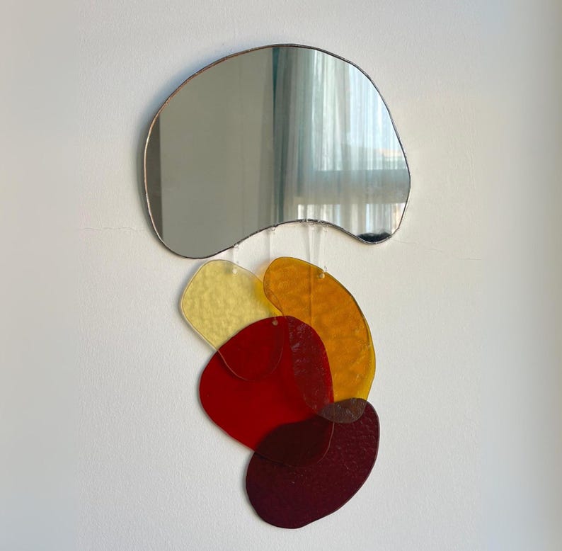 May include: A contemporary wall mirror with an unusual, organic form, framed in a thin metal. Beneath the mirror, a collection of colourful, translucent, abstract shapes in yellow, orange, and red are suspended.