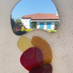 May include: An unusual wall mirror with a silver frame. The mirror reflects a house with a red tile roof and blue shutters. Below the mirror are layered, colourful, translucent shapes in shades of yellow, orange, and red.