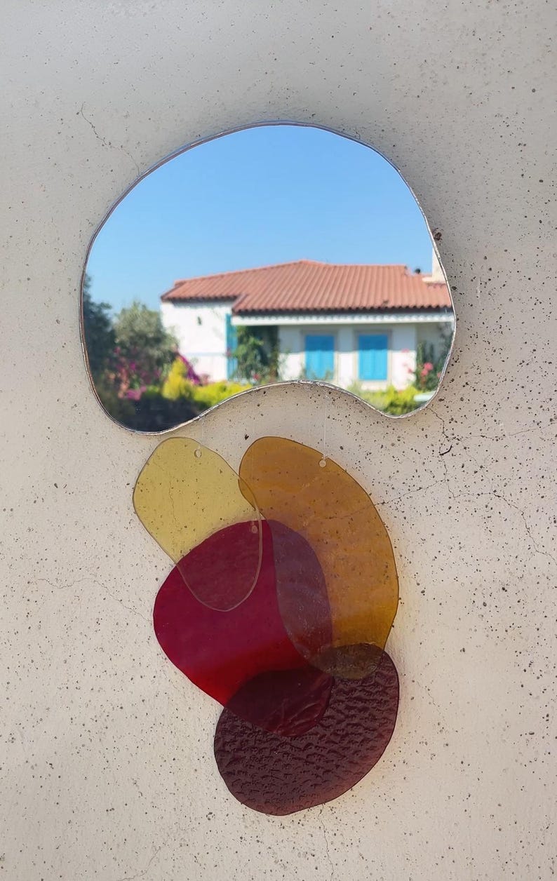 May include: An unusual wall mirror with a silver frame. The mirror reflects a house with a red tile roof and blue shutters. Below the mirror are layered, colourful, translucent shapes in shades of yellow, orange, and red.