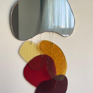 May include: Abstract wall art comprising a freeform mirror with a bronze-coloured frame above a cluster of colourful, irregularly shaped acrylic pieces. The acrylic shapes are in shades of yellow, orange, red, and burgundy, creating a layered, artistic composition.