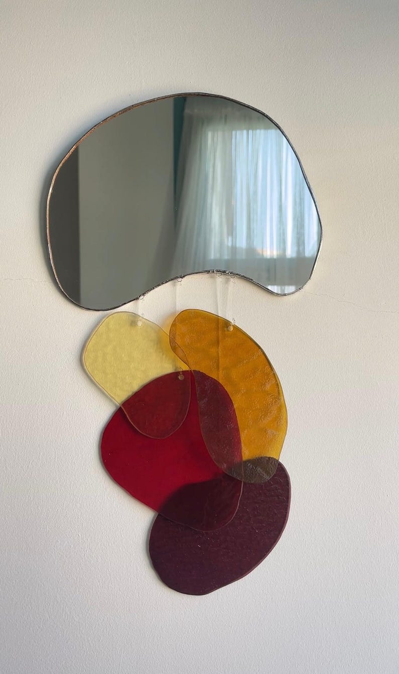 May include: Abstract wall art comprising a freeform mirror with a bronze-coloured frame above a cluster of colourful, irregularly shaped acrylic pieces. The acrylic shapes are in shades of yellow, orange, red, and burgundy, creating a layered, artistic composition.