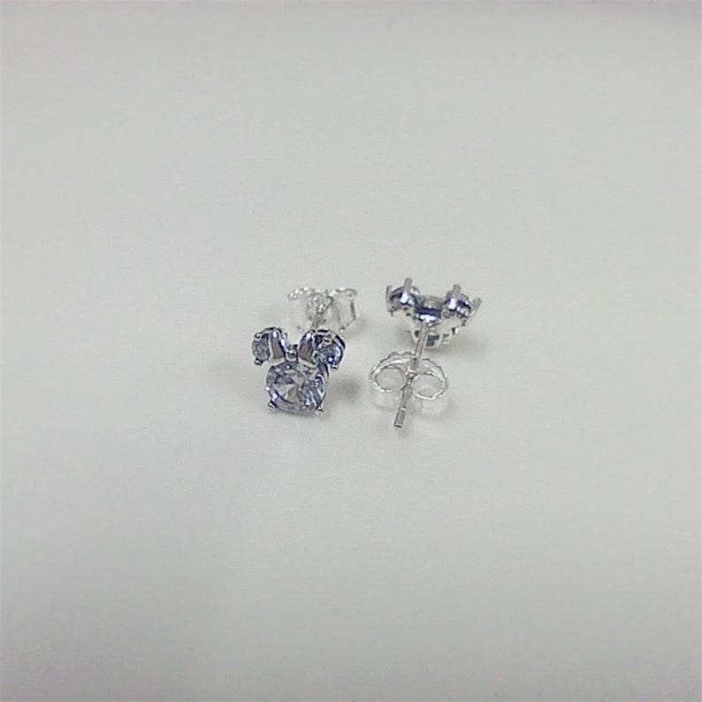May include: A pair of silver-colored stud earrings. Each earring features a three-part design, with a large, round, light blue gemstone at the center and two smaller gemstones above, resembling a bow. The earrings are on a white surface.