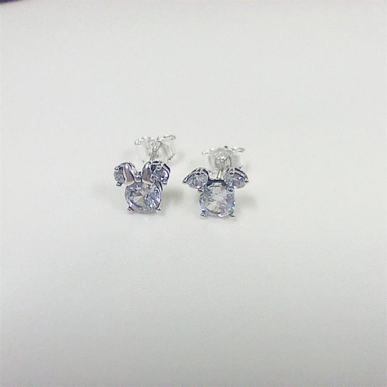 May include: A pair of silver-colored stud earrings. Each earring features a large, round, clear gemstone at the center, surrounded by smaller clear gemstones arranged in a bow shape. The earrings are on a white surface.