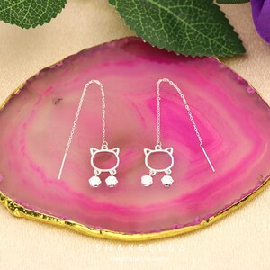 Puede incluir: Un par de pendientes colgantes plateados con forma de gato. Cada pendiente presenta una silueta de cabeza de gato con colgantes con forma de pata. Los pendientes se muestran sobre una superficie rosa y dorada, con hojas verdes y una flor morada en el fondo.