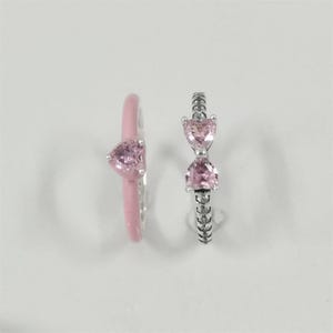 May include: Two rings with pink heart-shaped gemstones. One ring is silver with two heart-shaped stones. The other ring is pink with a single heart-shaped stone. The rings are set against a white background.
