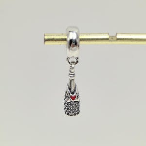 Puede incluir: Un charm de plata con un diseño de botella de champán. La botella tiene una apariencia texturizada, similar a una gema, con un detalle de corazón rojo. El charm está suspendido de un anillo de plata, ideal para pulseras o collares.