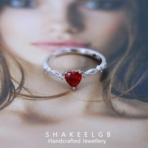 May include: A silver ring with a heart-shaped, deep red gemstone. The ring has a delicate design with small, clear accent stones. The brand name "SHAKEELGB Handcrafted Jewellery" is visible at the bottom.