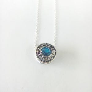 May include: A silver pendant necklace with a circular design. The pendant has a blue centre surrounded by concentric geometric patterns. The necklace chain is delicate and silver, creating a minimalist aesthetic.