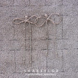 May include: Silver bow earrings with beaded chains. The earrings have a bow design at the top, with a small ball in the centre. The chains are decorated with small, evenly spaced beads. The text "SHAKEELGB Handcrafted Jewellery" is at the bottom.