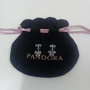 May include: A pair of silver stud earrings with a clear, square-cut gemstone. The earrings are displayed on a black velvet pouch with the brand name "PANDORA" printed in white. The pouch has a pink drawstring.